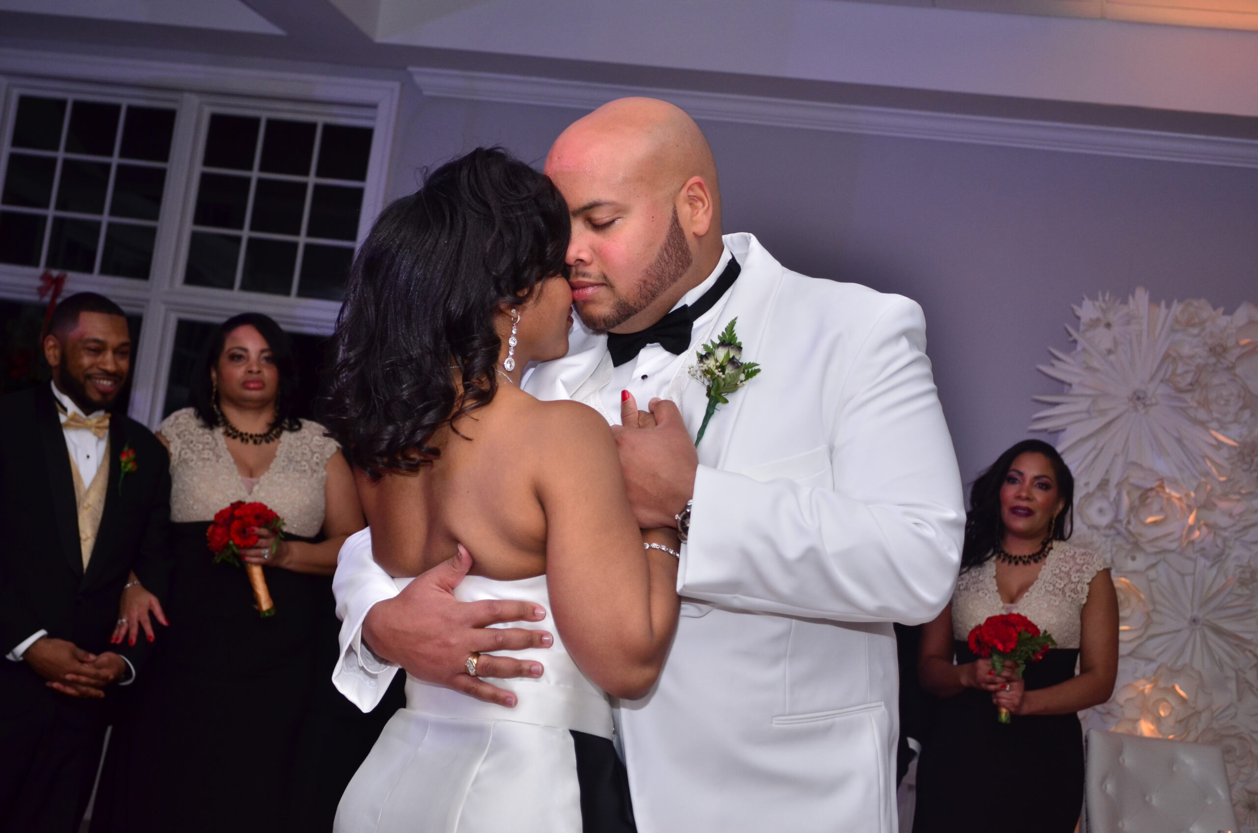 Bride and groom embrace in a close, intimate hug during their wedding reception, both in white formal wear with guests watching in the background.