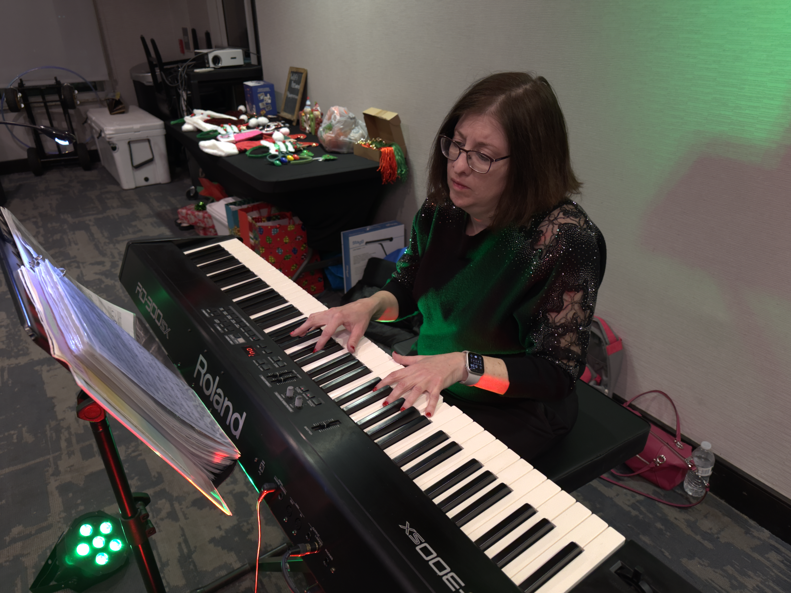 Woman in glasses playing a Roland keyboard in a cluttered room with a table of holiday gifts and equipment behind her.