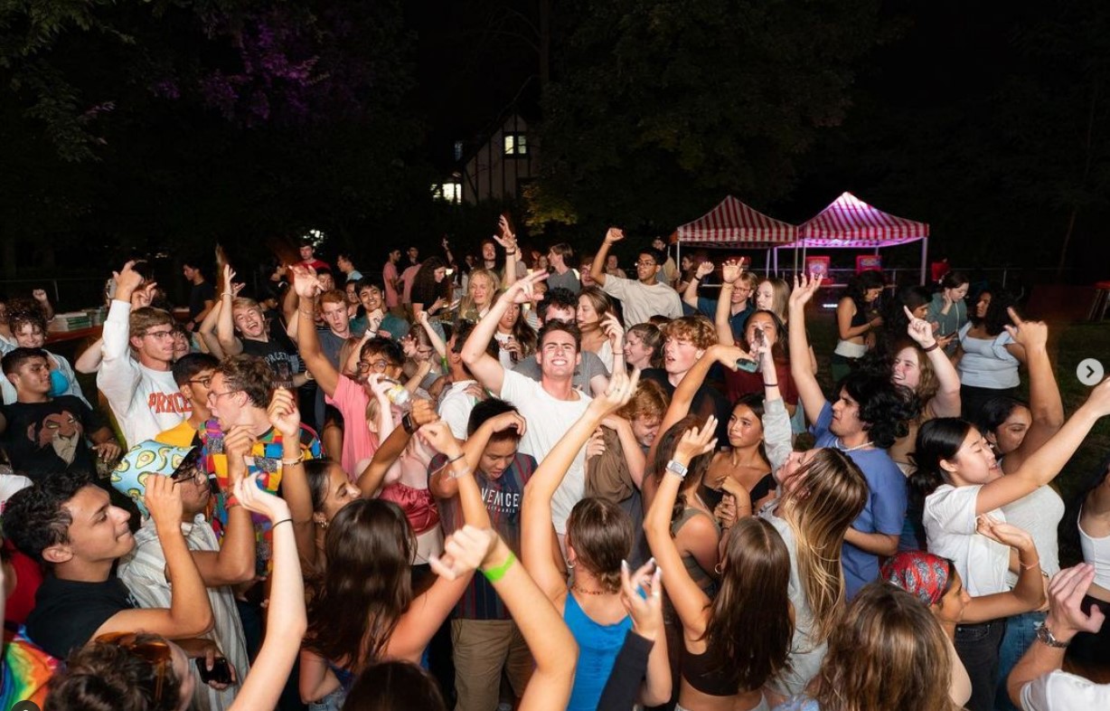 Large crowd of young people dancing and raising their hands at an outdoor night party, with striped tents in the background.