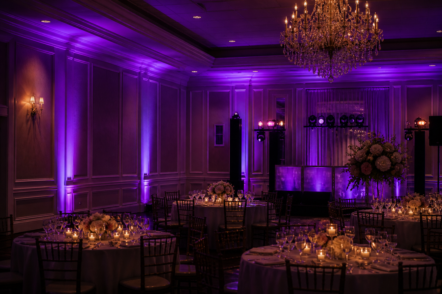 Elegant banquet hall with purple uplighting, a crystal chandelier, and round tables set with candles and white floral centerpieces. A DJ stage glows in the back.
