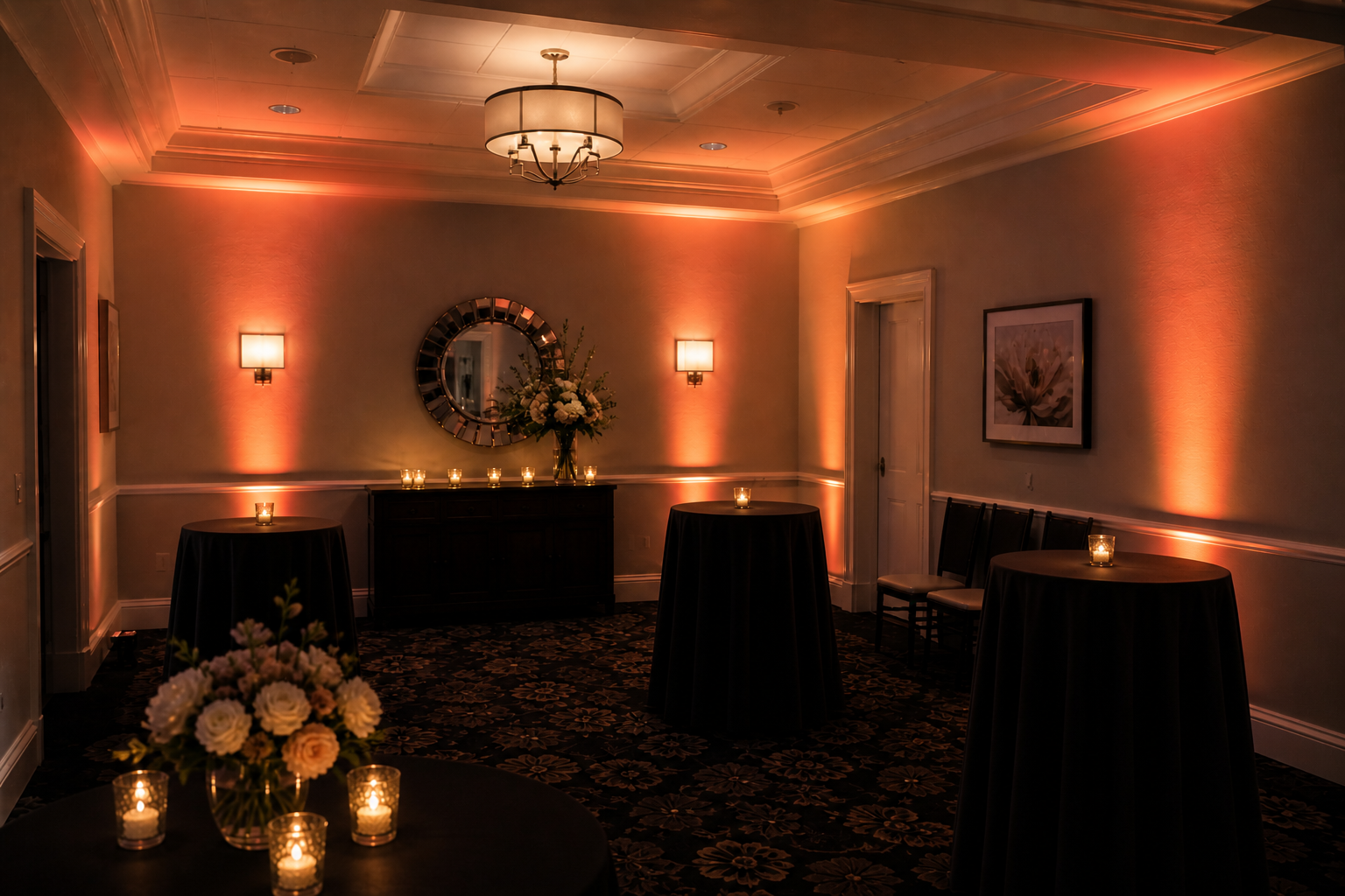 Dimly lit event room with round high-top tables covered in black cloth, candles, and a floral centerpiece on a dark sideboard.