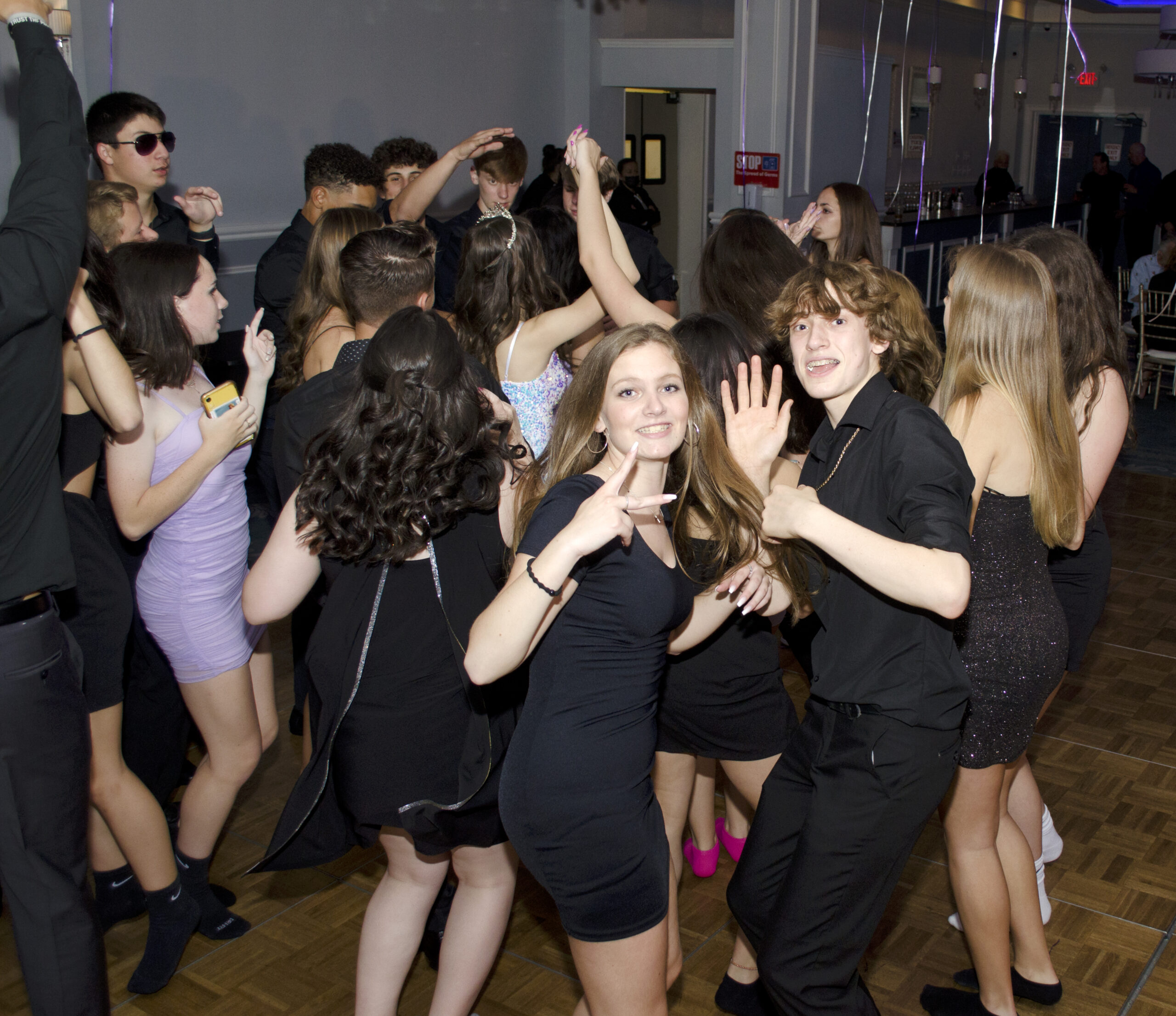 Group of teens dancing closely at a party, many raising hands and smiling in black outfits with balloons in the background.